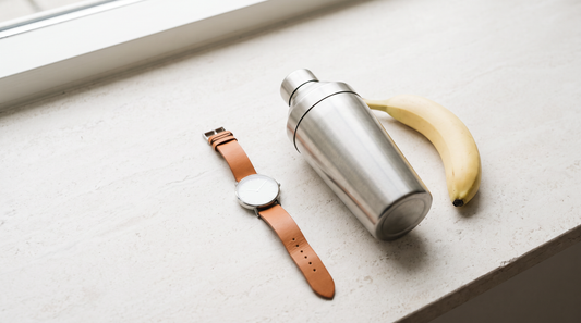 Armbanduhr, Shaker und Banane von oben auf heller Fläche als Sinnbild für Protein-Timing rund ums Training