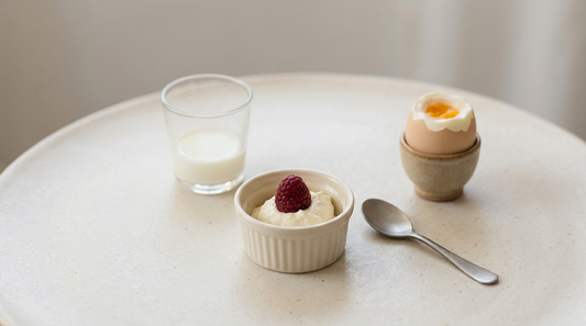 Drei kleine Portionen auf heller Fläche: ein Glas Milch, eine Schale Quark mit einer Beere und ein wachsweiches Ei im Becher